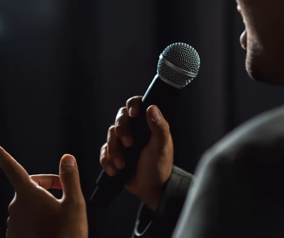 A person in low light holding a handheld microphone up to speak, seen over their shoulder