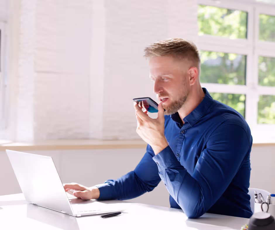 A man at a laptop in a bright office speaking into a phone held close to his mouth