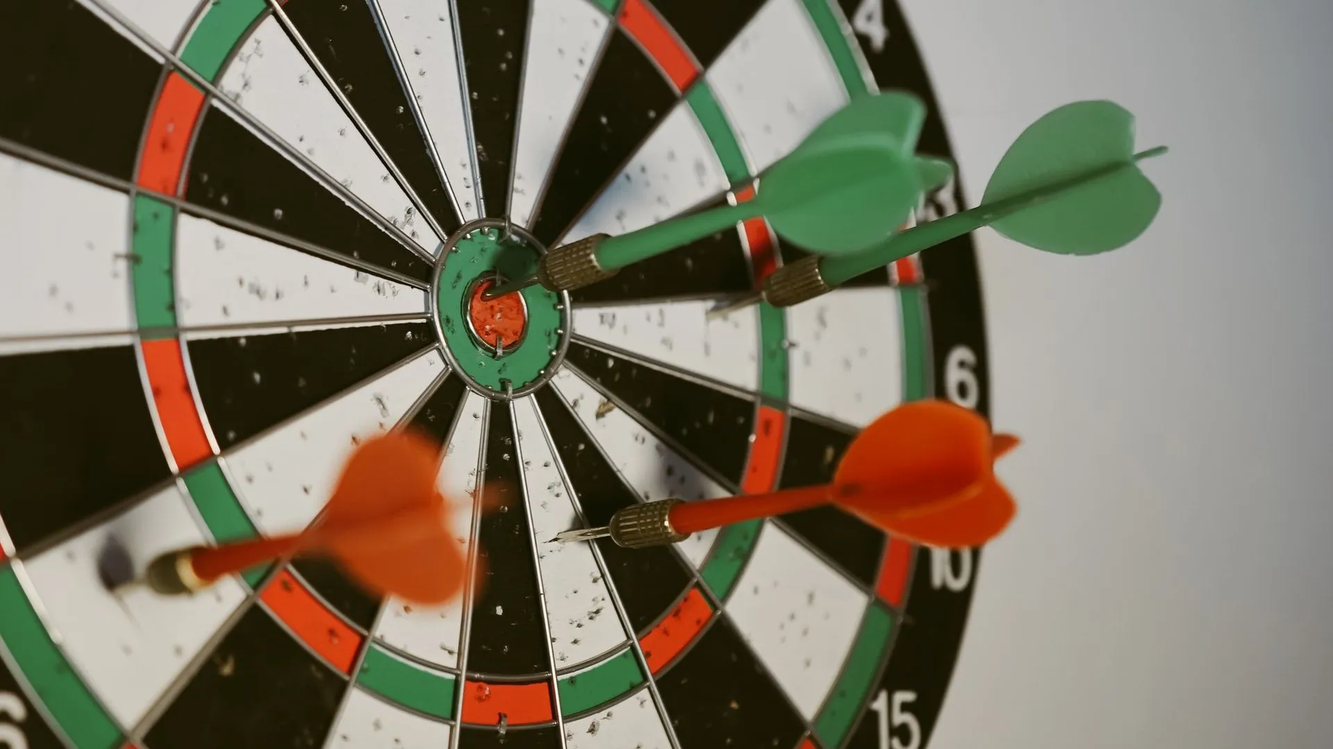 Close-up of a dartboard with a green dart in the bullseye and two orange darts nearby