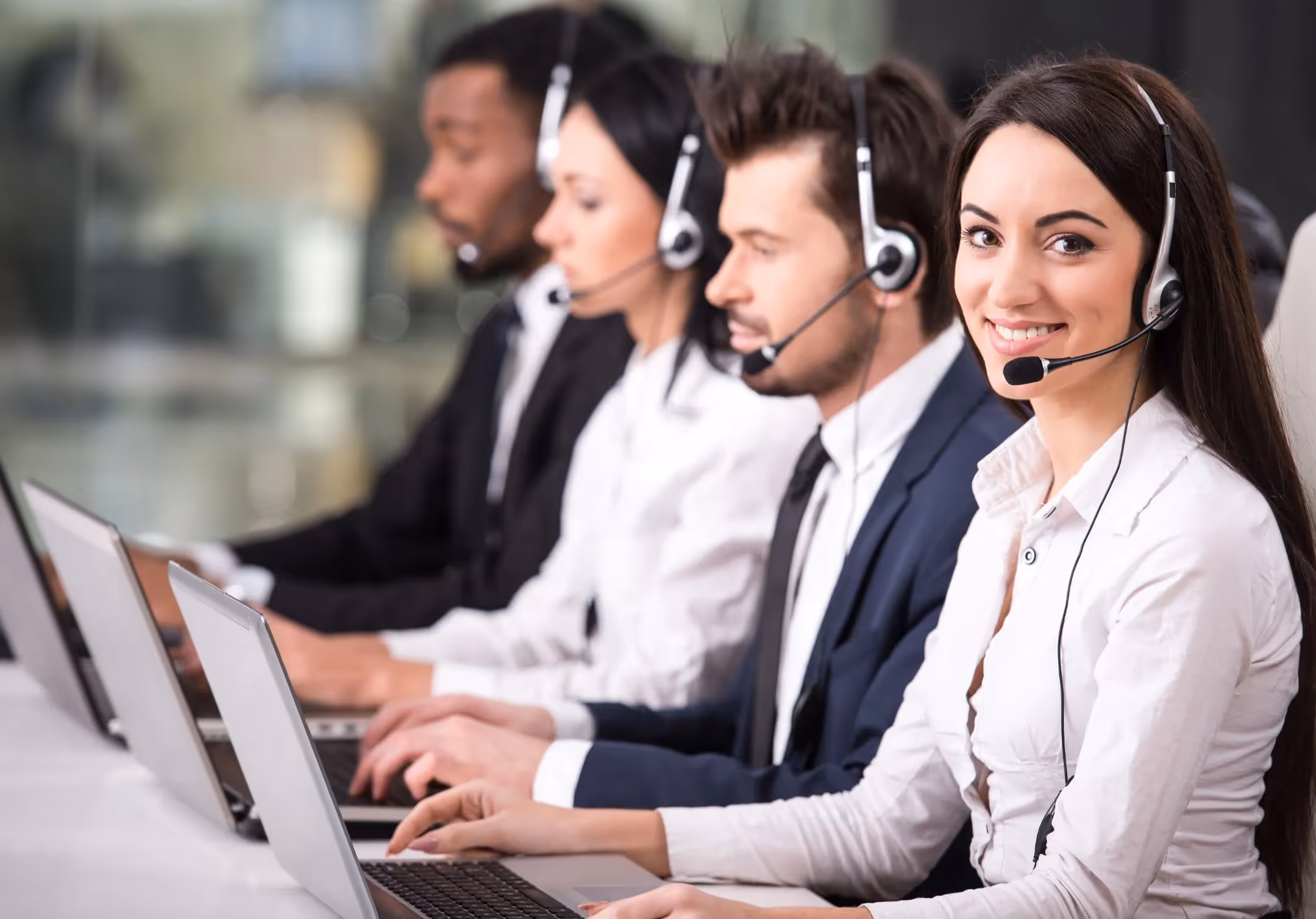 Four call center agents in headsets working at laptops in a row, the nearest smiling at the camera