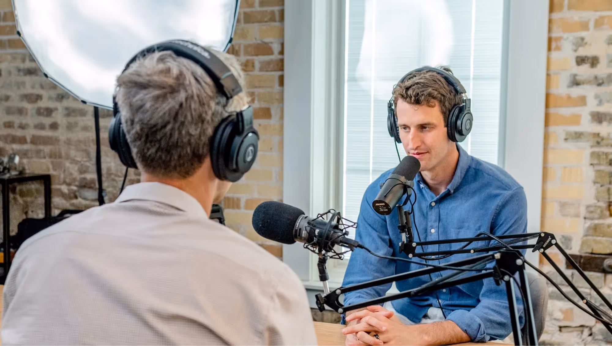 Two men in headphones facing each other across studio microphones in a brick-walled room